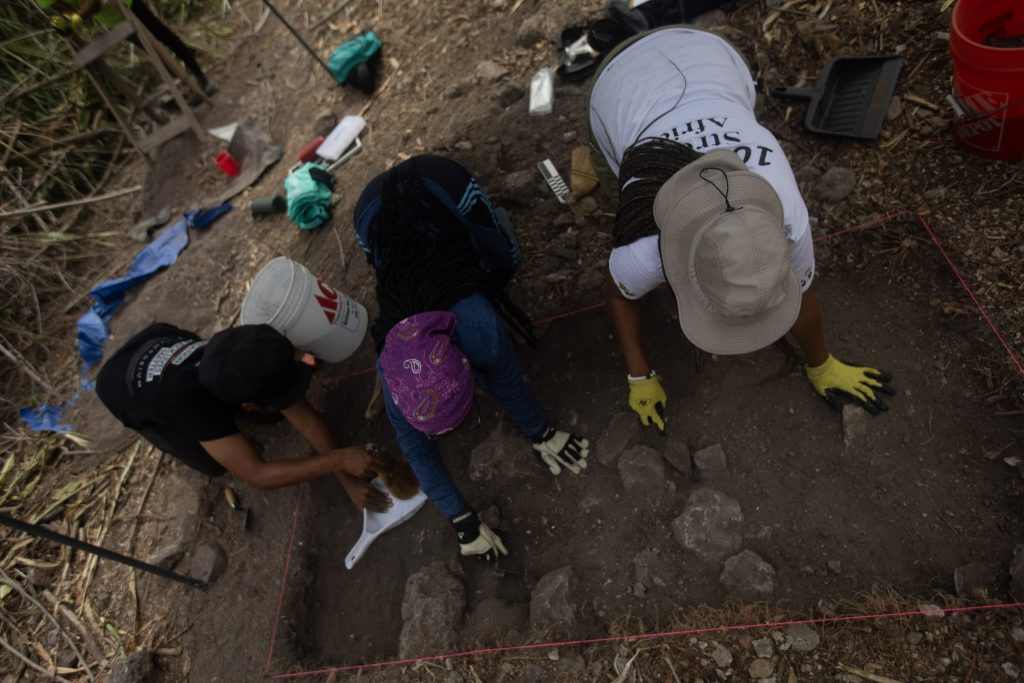 Three African American archaeologists are are on their hands and knees, excavating into the ground with tools.